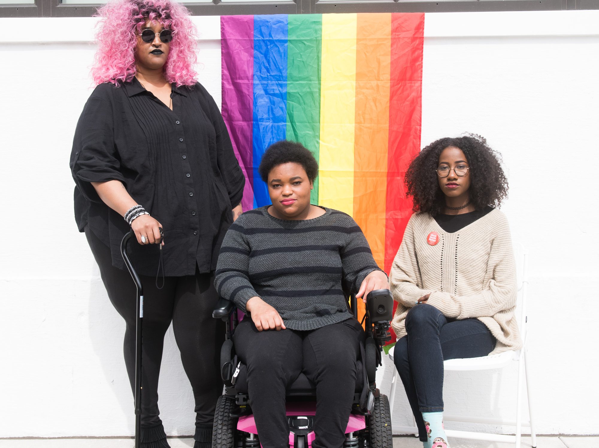 The Margins: Three Black and disabled folx (a non-binary person holding a cane, a woman sitting in a power wheelchair, and a woman sitting in a chair) looking seriously at the camera while a rainbow pride flag drapes on the wall behind them. Credit: Disabled and Here